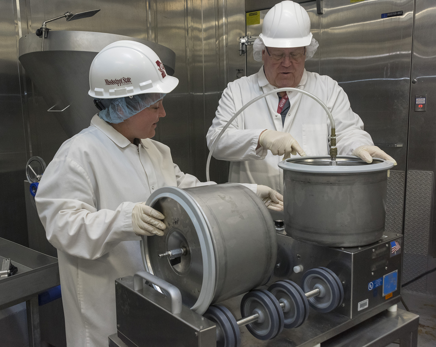 A woman and a man wearing lab coats, hard hats, and hair nets operating equipment in a lab.