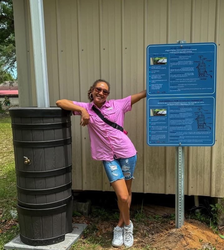 Woman standing next to a rain barrel smiling with sunglasses on.