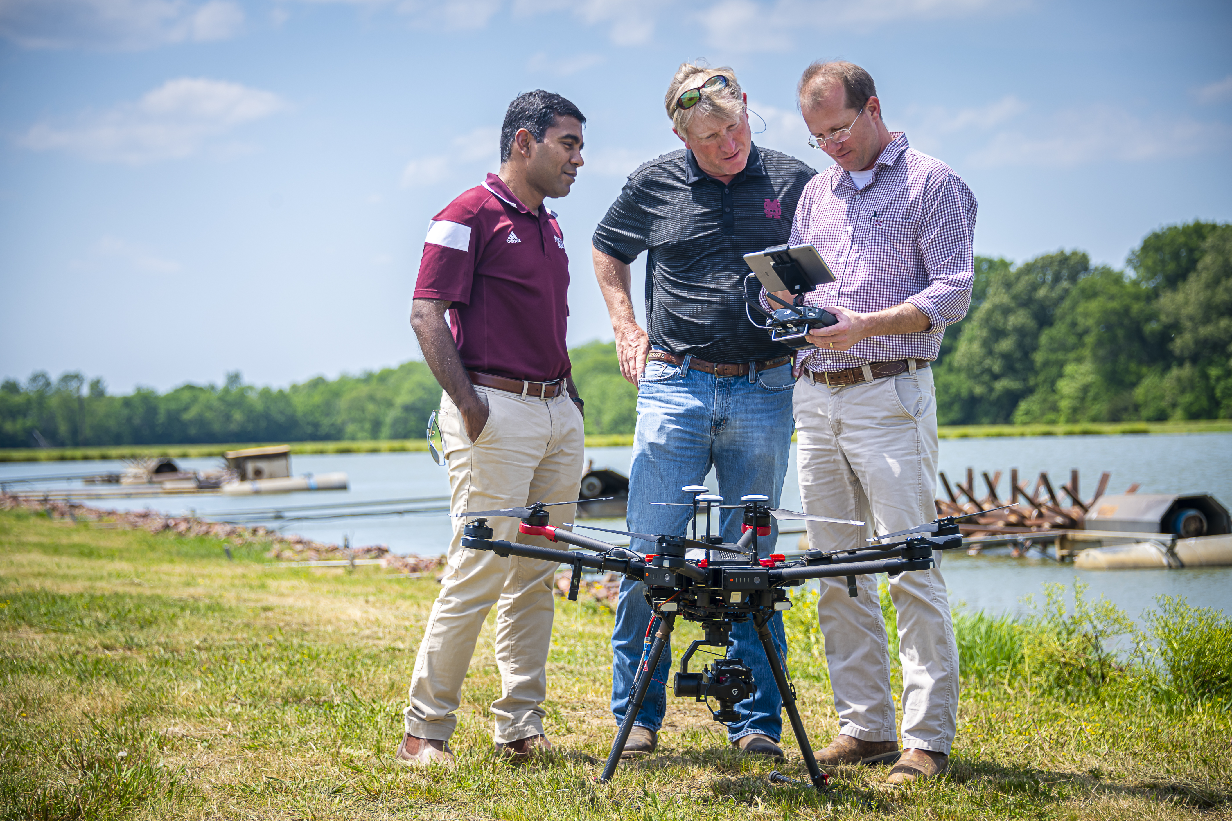 Three man standing in front of a catfish pond next to a drone sitting on the ground, looking at an ipad. 